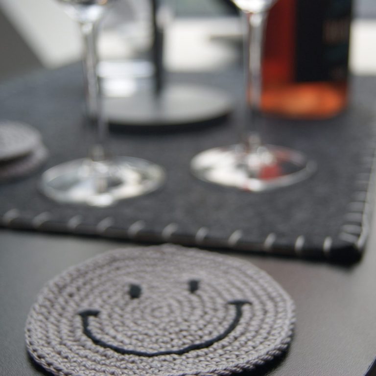Untersetzer mit einem Smilie-Gesicht, im Hintergrund sind Weingläser, eine Flasche und eine Lampe zu sehen.